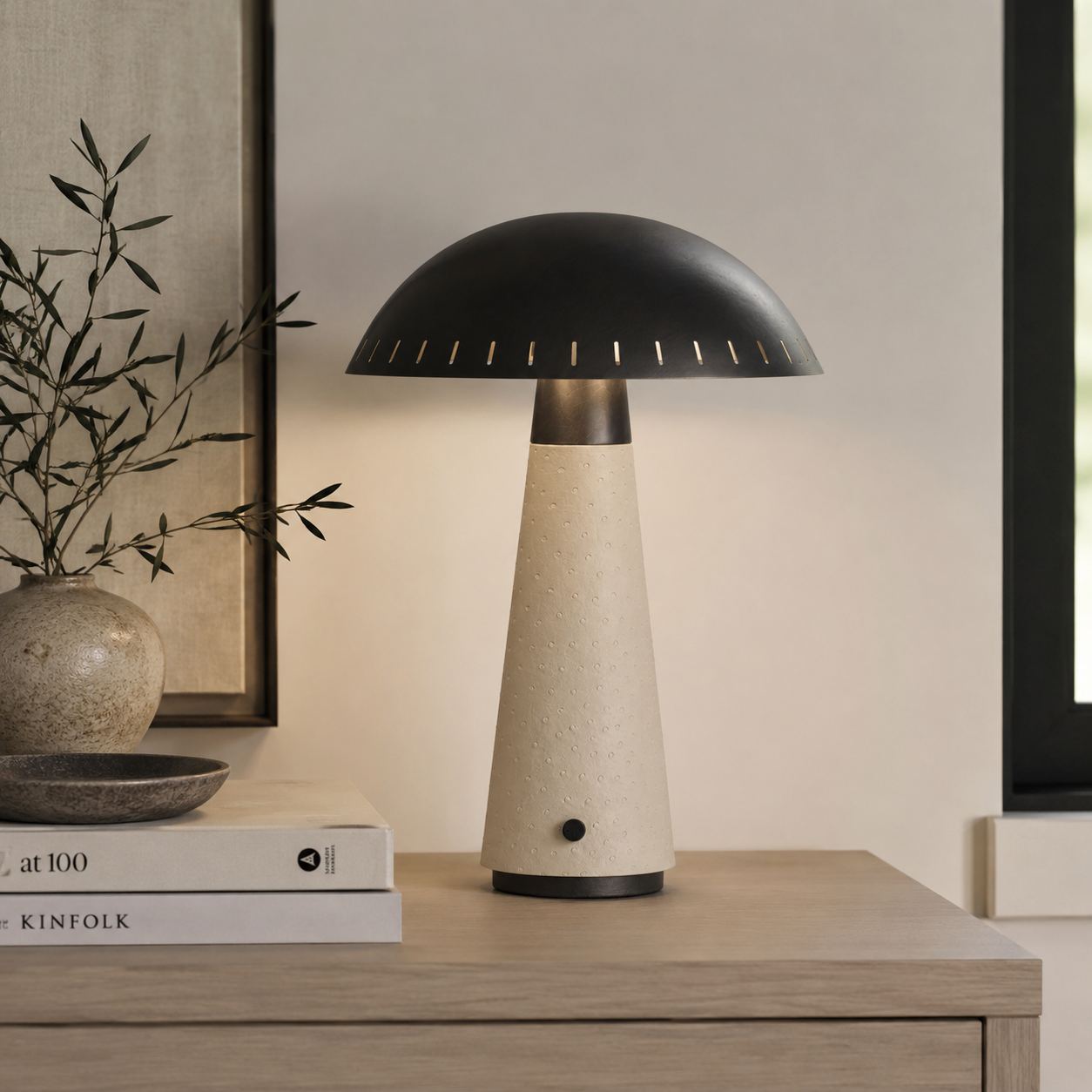 A mushroom-shaped table lamp with a matte black metal dome shade and a textured beige stone-like base, styled on a light wood sideboard next to a vase and books.