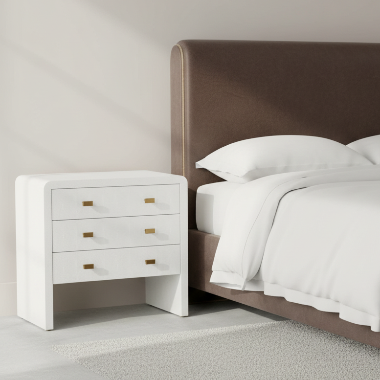 White nightstand with gold handles next to a bed with white bedding and a brown headboard. harlow