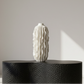 White textured vase on a black textured pedestal against a neutral wall.