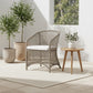Wicker chair with a white cushion on a patio with potted plants and a wooden side table.
