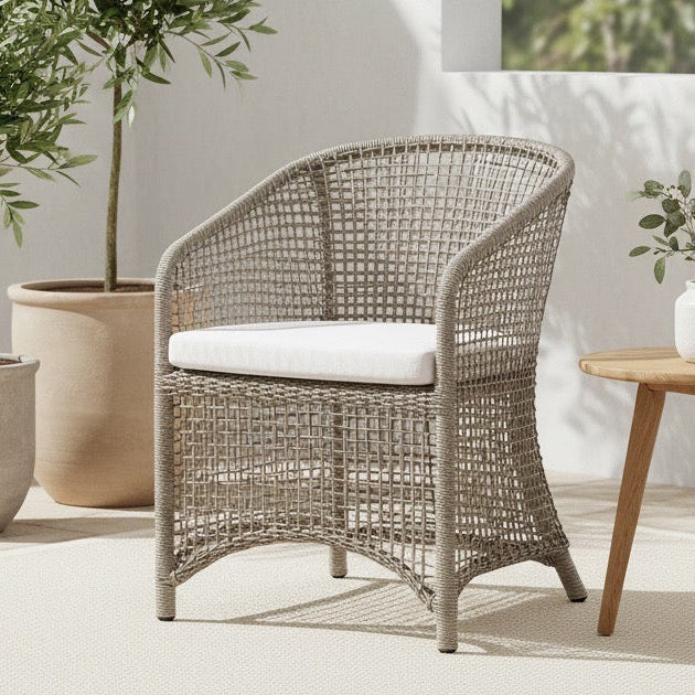 Wicker chair with a white cushion on a patio with potted plants and a wooden side table.