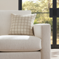 Beige sofa with a checkered pillow in a room with large windows showing greenery outside.