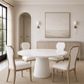 A round white dining table with four upholstered chairs in a bright, modern dining room. The room features a light beige sideboard, a large abstract painting, gold wall sconces, and a black-framed window on the left.