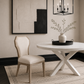 A styled dining room scene featuring a white round table with a white X-base, a light beige upholstered dining chair with a light wood frame, a white ceramic vase with dried branches on a black tray, and a black wall-mounted candelabra hanging above.
