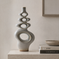 A tall, sculptural ceramic vase with stacked abstract oval and circular cutouts, finished in a matte light gray glaze with subtle gold and brown speckled detailing near the base and openings. It stands on a stone ledge beside stacked books and a small bowl against a warm beige wall.