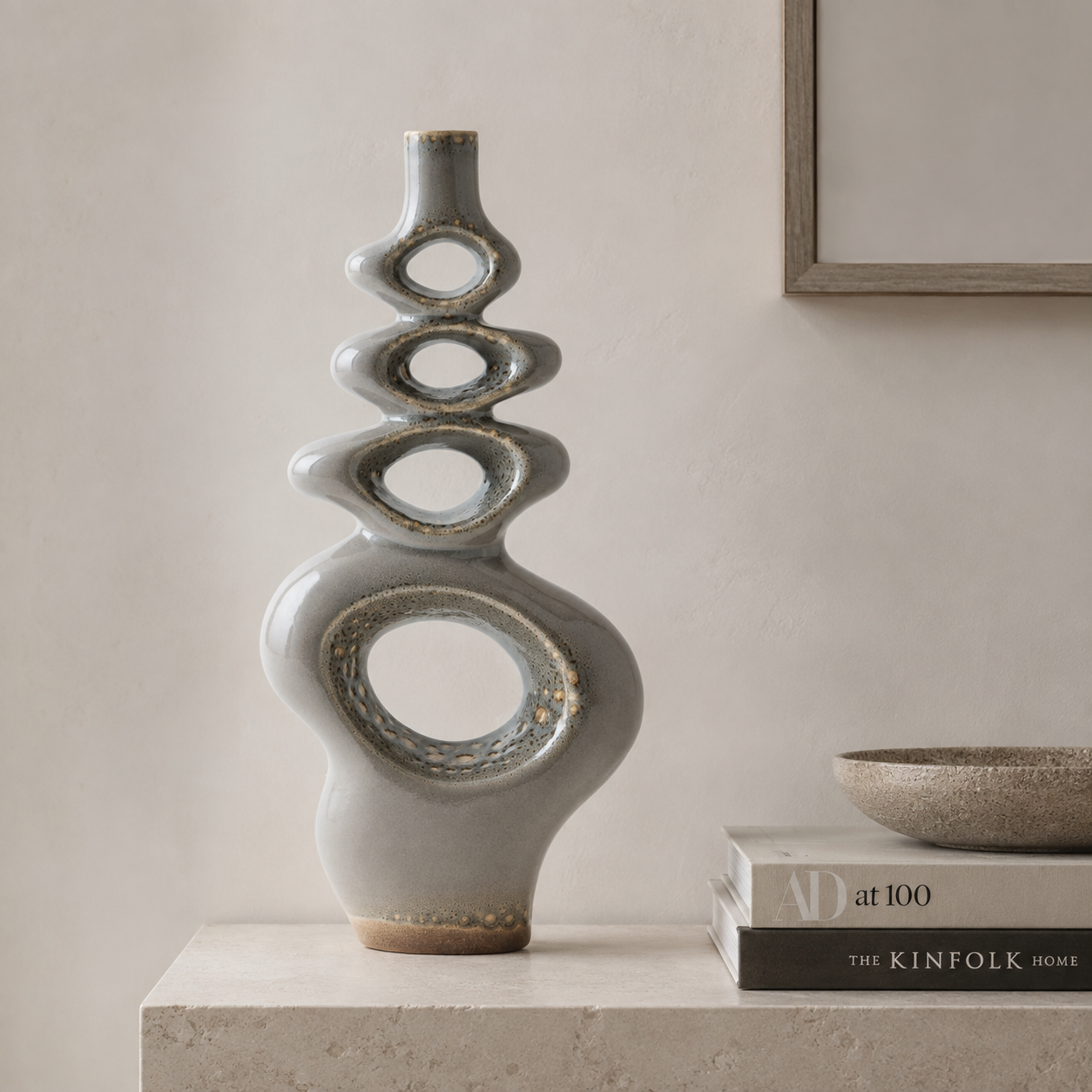 A tall, sculptural ceramic vase with stacked abstract oval and circular cutouts, finished in a matte light gray glaze with subtle gold and brown speckled detailing near the base and openings. It stands on a stone ledge beside stacked books and a small bowl against a warm beige wall.