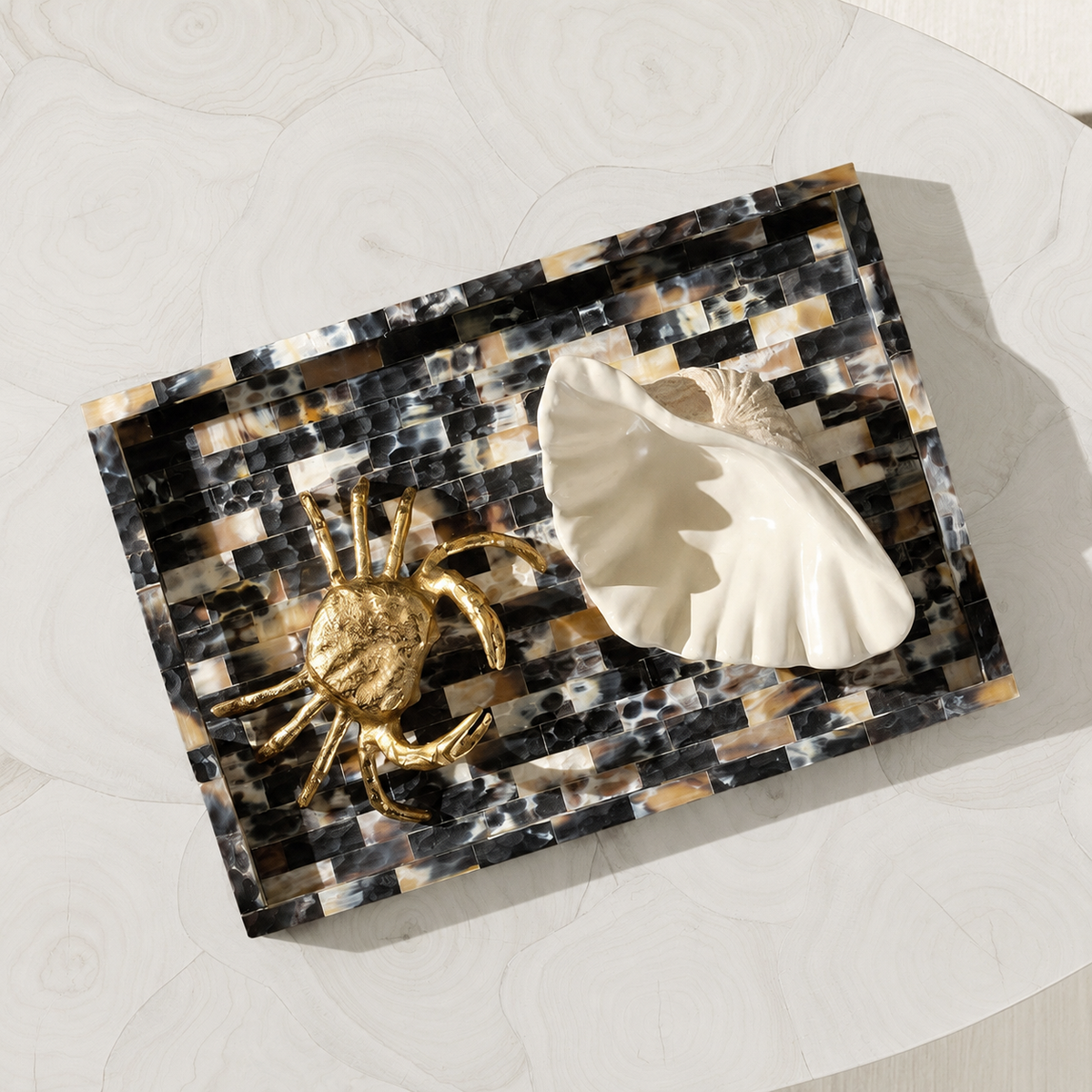 A rectangular decorative tray with a black, white, and beige marbled mosaic pattern, styled with a gold crab figurine and a white ceramic seashell on a white marble surface.