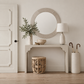 A styled entryway scene featuring a white console table beneath a round decorative mirror, with a vase of greenery, a table lamp, a carved wooden stool, and a cream umbrella holder on a light wood floor.