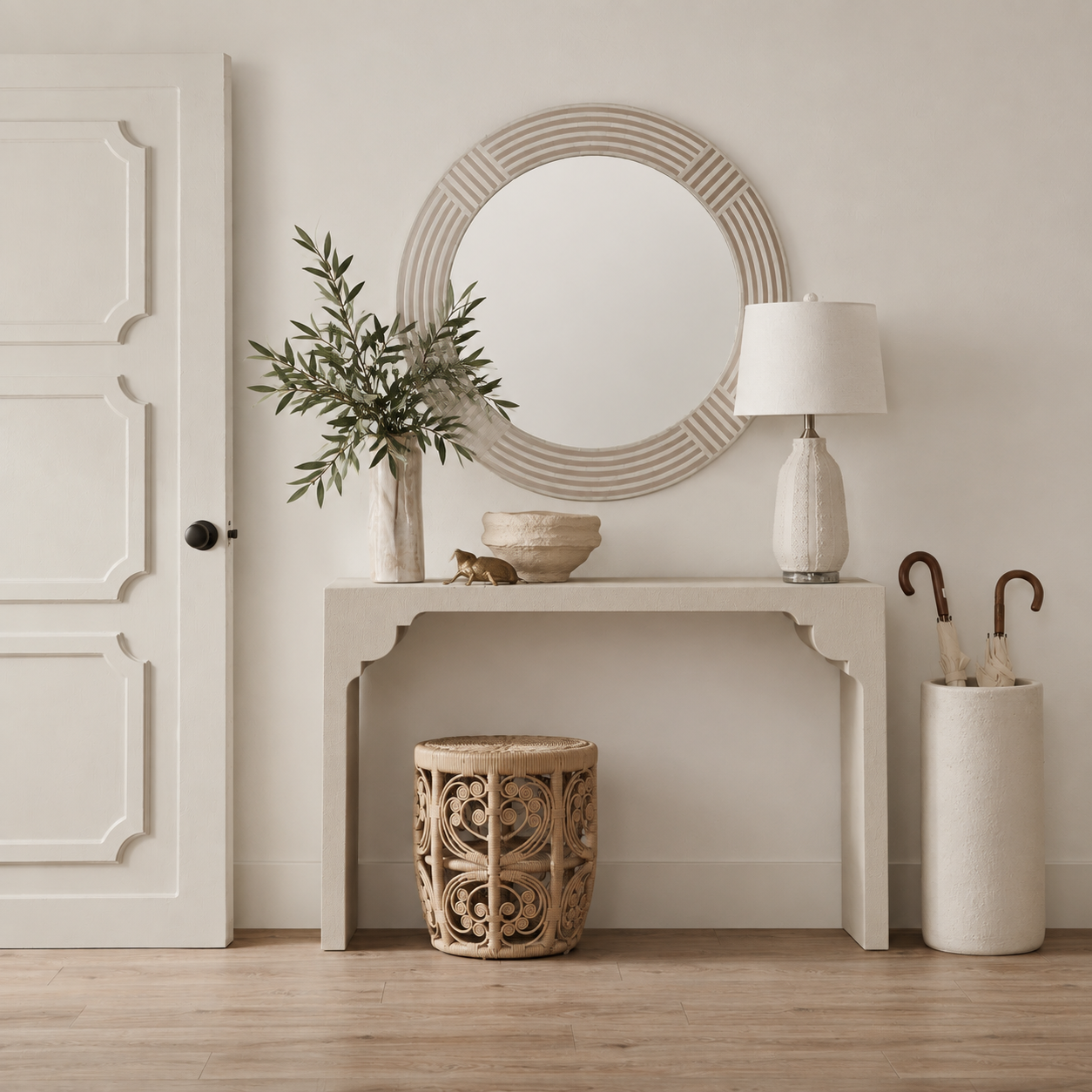 A styled entryway scene featuring a white console table beneath a round decorative mirror, with a vase of greenery, a table lamp, a carved wooden stool, and a cream umbrella holder on a light wood floor.