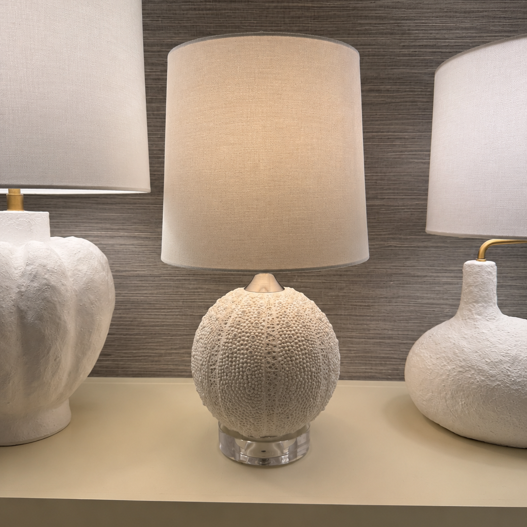 A table lamp with a textured, spherical sea urchin-inspired base and a cylindrical beige linen shade, displayed on a shelf between other decorative lamps against a gray textured wall.