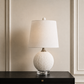 A table lamp with a textured white spherical base and a tapered off-white fabric shade, set on a dark wooden surface against a beige paneled wall.