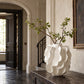 Sculptural ivory ceramic vase with dramatic flowing ruffled details, styled with leafy green branches on a dark wood console table in an elegant French-inspired entryway featuring textured neutral walls, an arched doorway, and classic architectural details.
