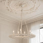 A large, ornate white chandelier with a leaf-like circular frame and multiple candle-style bulbs, hanging from chains in a decorated room with a detailed white ceiling medallion and curtains.