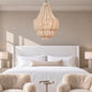 A large, tiered bohemian-style chandelier made of woven natural fiber hangs over a neatly made bed in a modern, neutral-toned bedroom with two fluffy armchairs in the foreground.