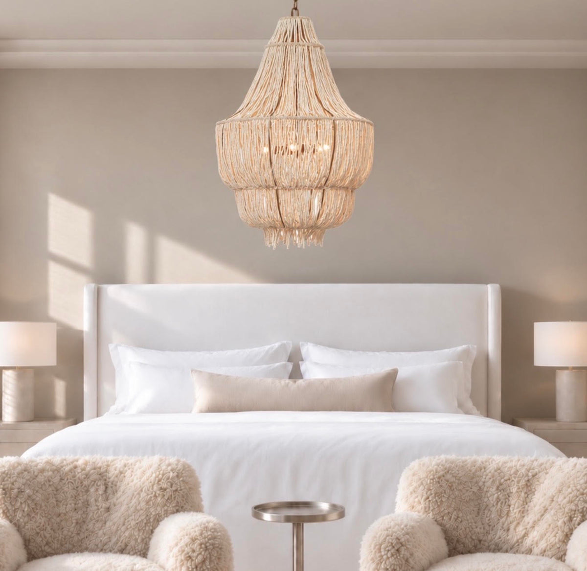 A large, tiered bohemian-style chandelier made of woven natural fiber hangs over a neatly made bed in a modern, neutral-toned bedroom with two fluffy armchairs in the foreground.