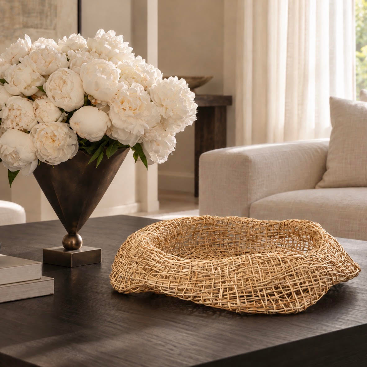 A natural woven rattan bowl or tray sits on a dark wooden coffee table in a modern living room setting, styled with a large vase of white peonies and books.