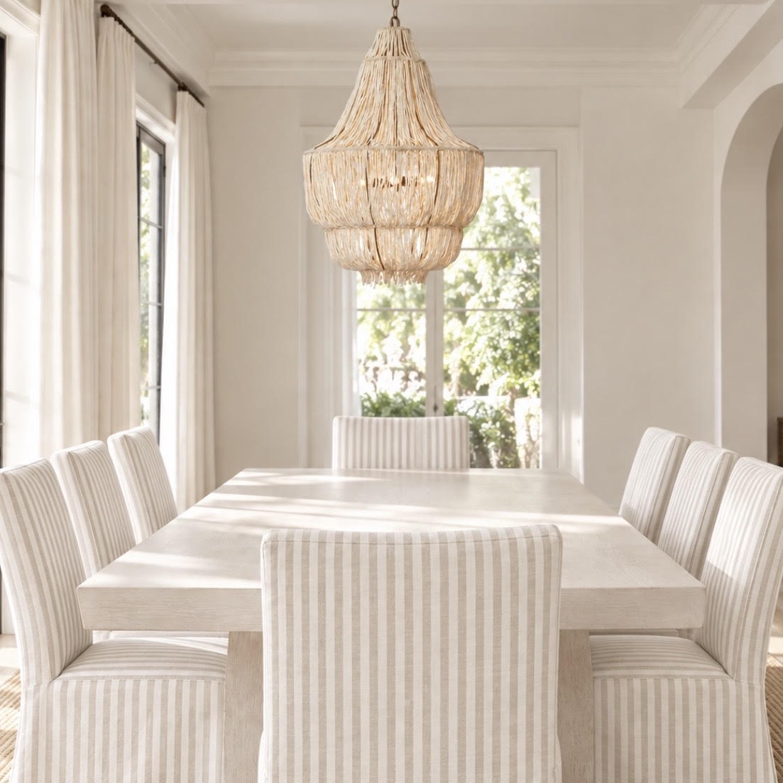 A bright dining room with a long white table and multiple upholstered chairs in a light beige and white stripe pattern, under a large woven pendant chandelier.