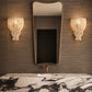A luxury bathroom scene featuring a large, asymmetrical wavy mirror with a gold frame, flanked by two woven macrame wall sconces. Below the mirror is a gold faucet and a white marble sink with dramatic black veining, all set against a dark textured wood-paneled wall.