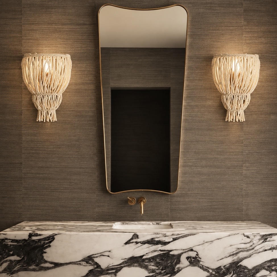 A luxury bathroom scene featuring a large, asymmetrical wavy mirror with a gold frame, flanked by two woven macrame wall sconces. Below the mirror is a gold faucet and a white marble sink with dramatic black veining, all set against a dark textured wood-paneled wall.