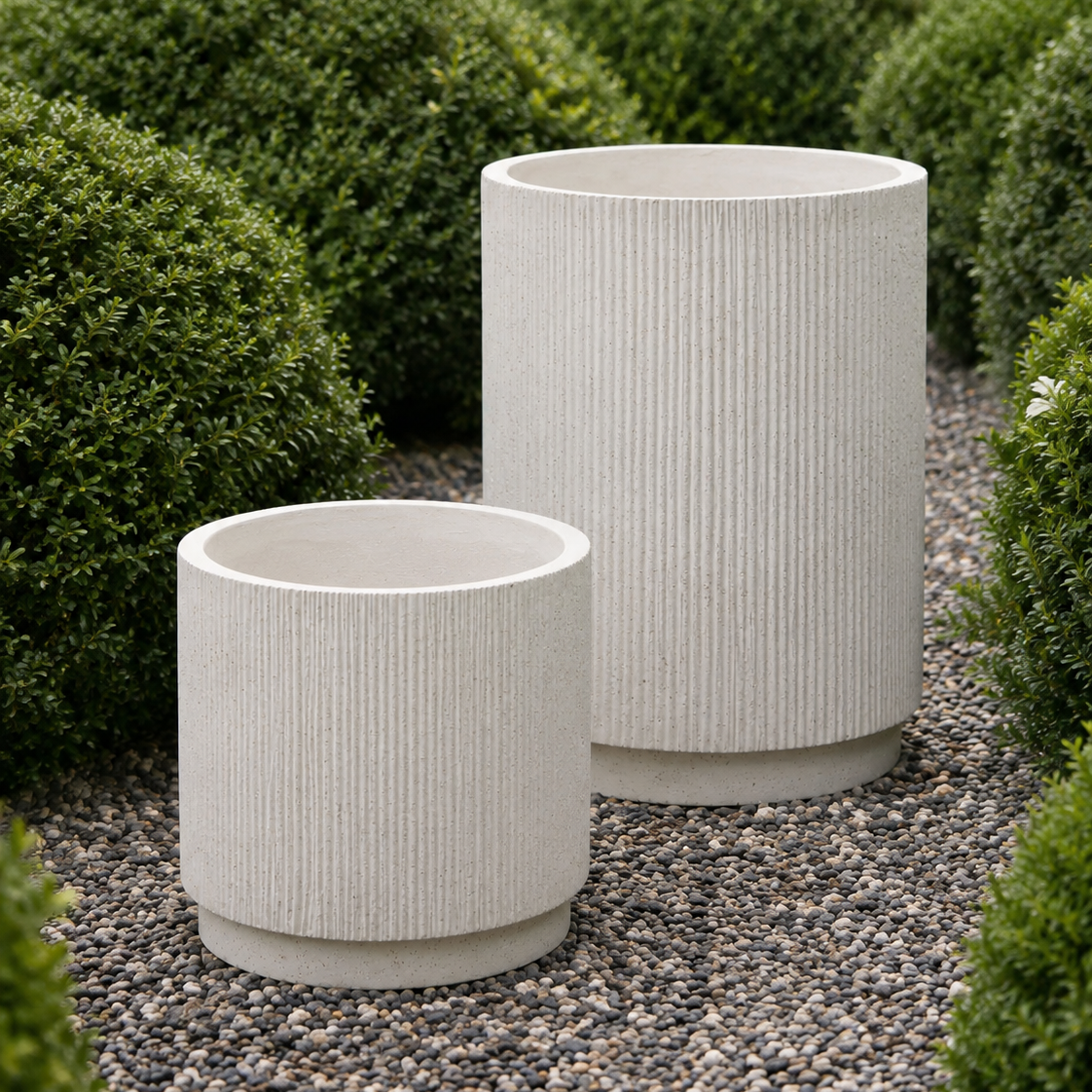 Two ribbed cylindrical planters in a light warm gray or off-white stone-like finish, placed on a gravel path surrounded by trimmed green bushes.