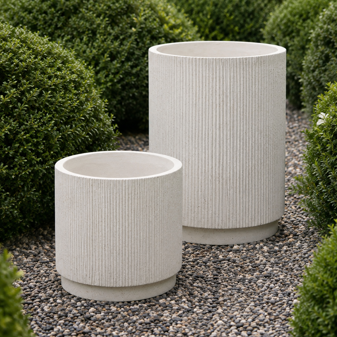 Two ribbed cylindrical planters in a light warm gray or off-white stone-like finish, placed on a gravel path surrounded by trimmed green shrubs.