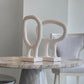 Two abstract sculptural tabletop decor pieces in a warm off-white/cream color, displayed on a round gray marble dining table next to a light gray upholstered chair.