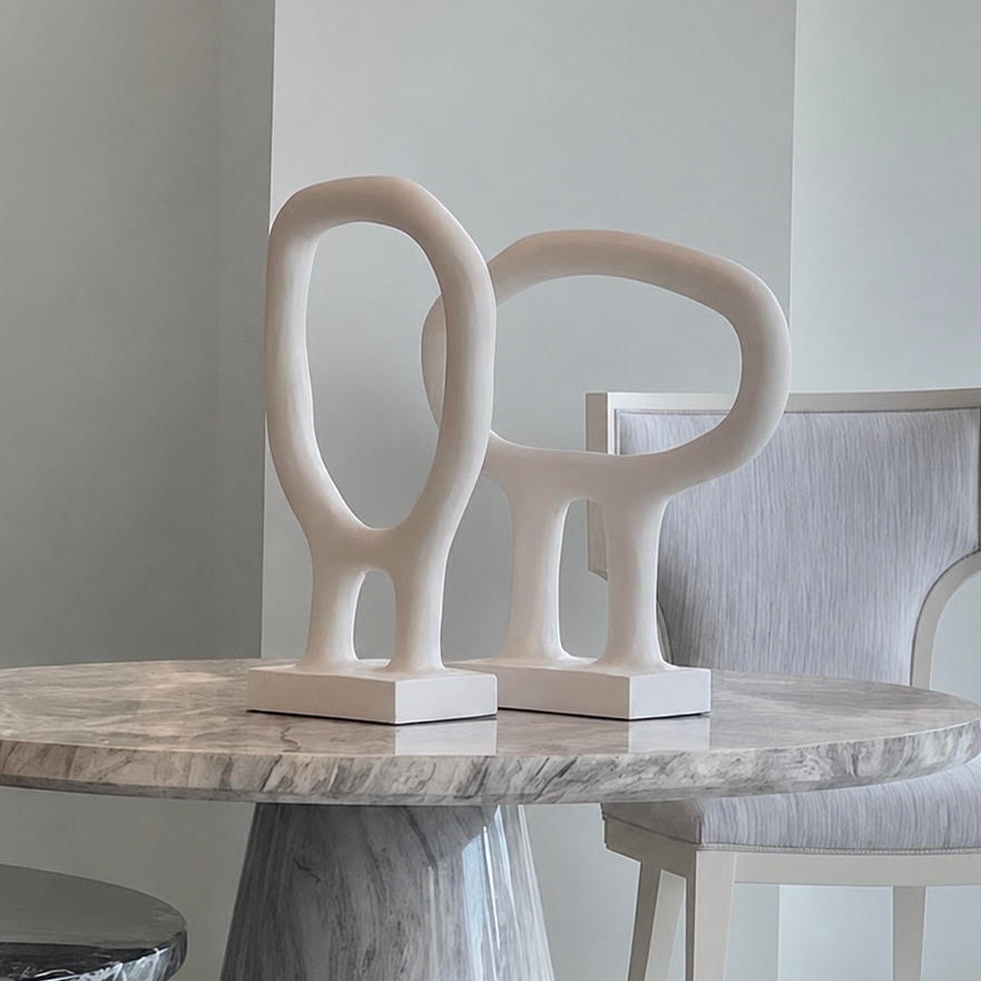 Two abstract sculptural tabletop decor pieces in a warm off-white/cream color, displayed on a round gray marble dining table next to a light gray upholstered chair.