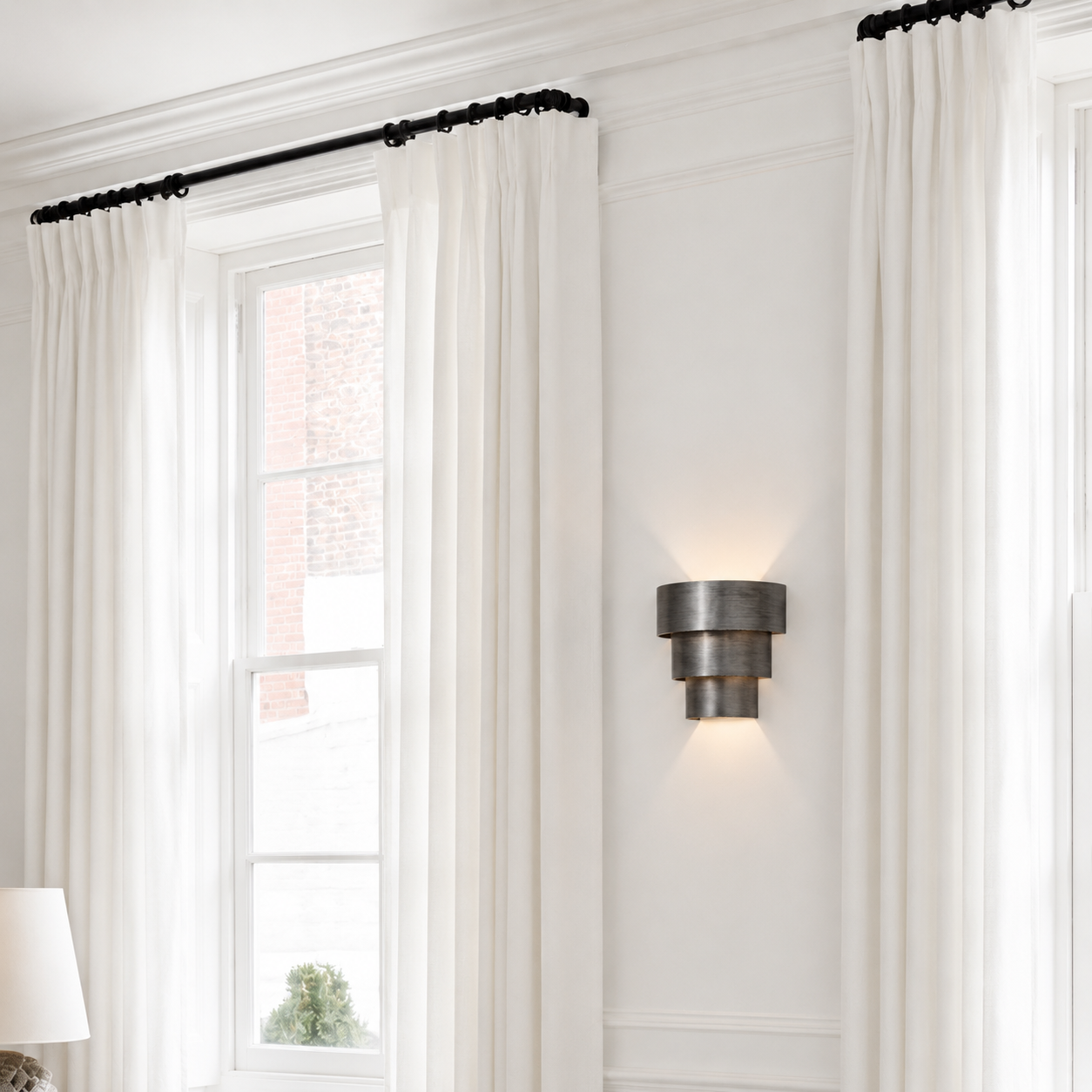 White curtain panels hanging from a black rod in a bright room with white walls and a wall-mounted light fixture.