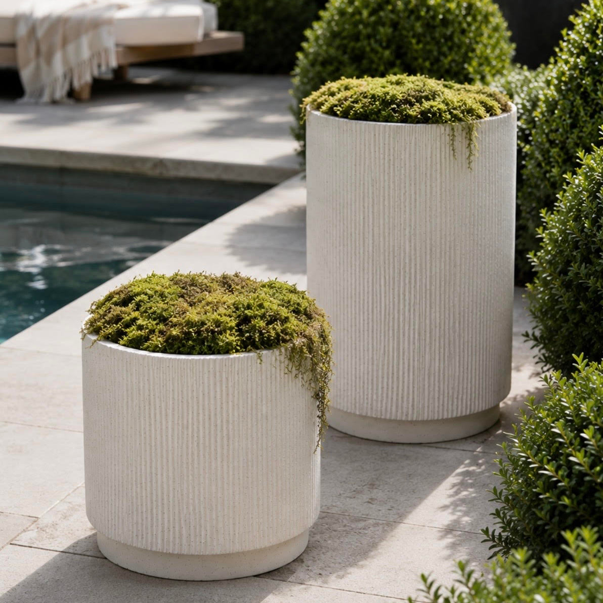Two tall ribbed white concrete planters filled with green moss, placed on a stone patio beside a swimming pool and landscaped shrubs.