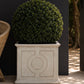 A round, dense green topiary tree planted in a large, square, light gray concrete planter with embossed geometric patterns, set against a dark charcoal brick wall.