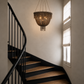 A rustic woven chandelier with hanging tassels is suspended in a staircase landing with dark wood steps, a black curved handrail, and tall windows with white blinds.