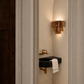 A modern wall-mounted bathroom sink with a black basin and brass faucet, set in a narrow alcove with a cylindrical wood-grain sconce on the wall. A white towel is draped over the sink, and the scene is framed by white door trim on the left and right.