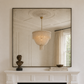 A large rectangular wall mirror with a thin dark frame hangs above a white fireplace mantel. The mirror reflects a cream chandelier, window, and curtains, while the mantel also holds a small bust sculpture and stacked books.