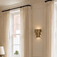 A pair of floor-length cream curtains hanging from a black rod over a tall window in a bright, elegant room with a gold wall sconce.
