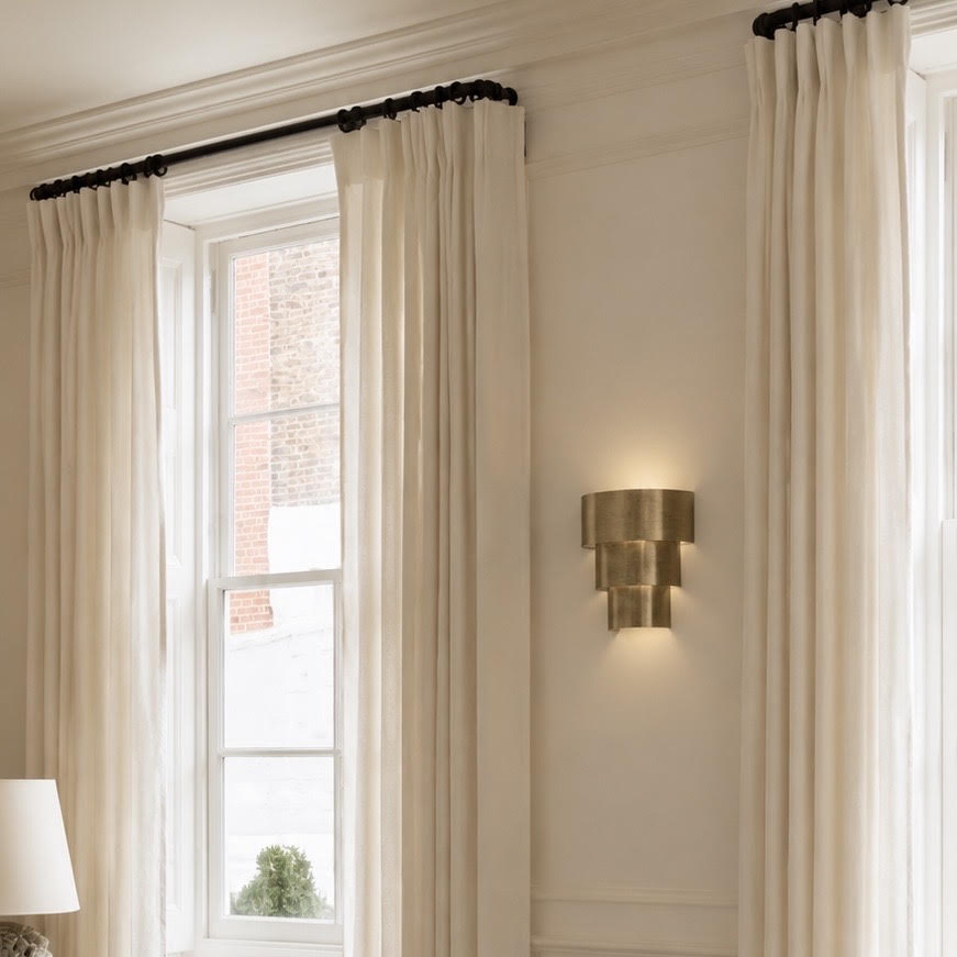 A pair of floor-length cream curtains hanging from a black rod over a tall window in a bright, elegant room with a gold wall sconce.