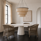 A modern dining room setup featuring a round white pedestal table surrounded by four woven wicker armchairs with white cushions, under a large tiered beige fabric chandelier.