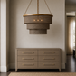 A modern tiered chandelier with a woven taupe fabric shade and gold-toned metal trim hangs above a matching six-drawer dresser in a neutral-toned room.