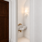 Elegant minimalist powder room featuring a sculptural white leaf-inspired wall sconce, floating white marble sink with integrated basin, antique brass wall-mounted faucet, arched alcove, classic white wall molding, and a rich dark wood door. Soft warm lighting creates a refined, modern French-inspired aesthetic.