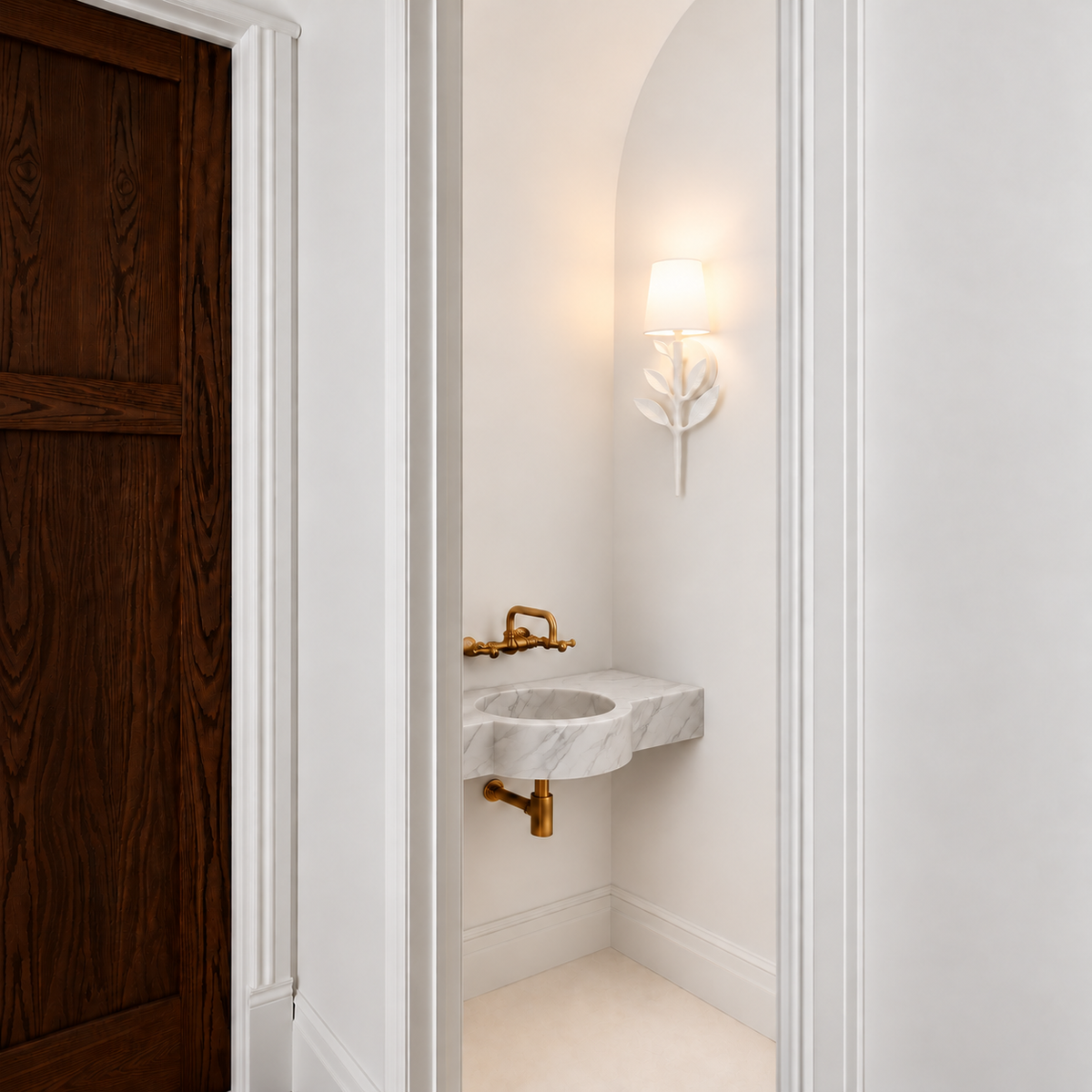 Elegant minimalist powder room featuring a sculptural white leaf-inspired wall sconce, floating white marble sink with integrated basin, antique brass wall-mounted faucet, arched alcove, classic white wall molding, and a rich dark wood door. Soft warm lighting creates a refined, modern French-inspired aesthetic.