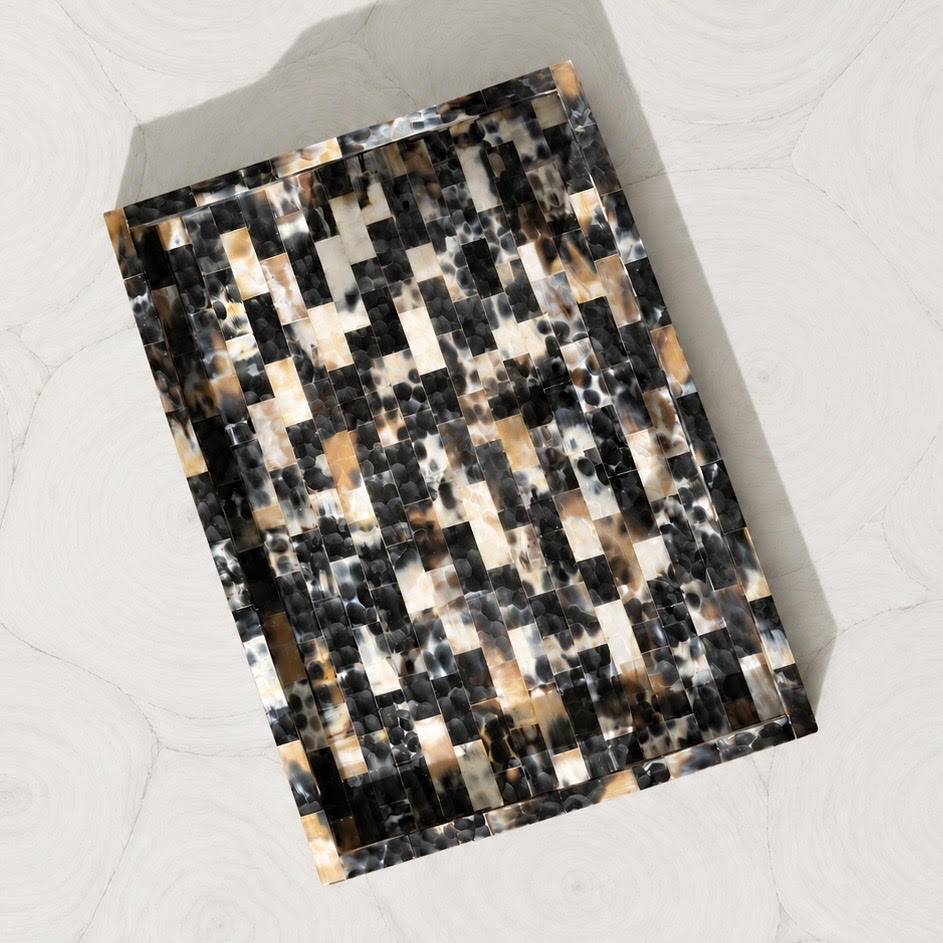 A rectangular tray with a mosaic tile pattern in black, white, and warm brown, photographed from an overhead angle on a light marble surface.