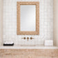 A rectangular wall mirror with a woven natural fiber border hangs above a sink in a bathroom with white tiled walls, brass faucet handles, a light stone countertop, and folded white towels.