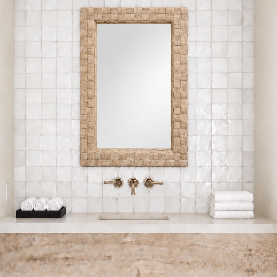 A rectangular wall mirror with a woven natural fiber border hangs above a sink in a bathroom with white tiled walls, brass faucet handles, a light stone countertop, and folded white towels.