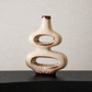 A modern, abstract ceramic vase with a glossy cream-colored glaze and brown speckled accents, displayed on a dark wooden surface against a textured beige wall.