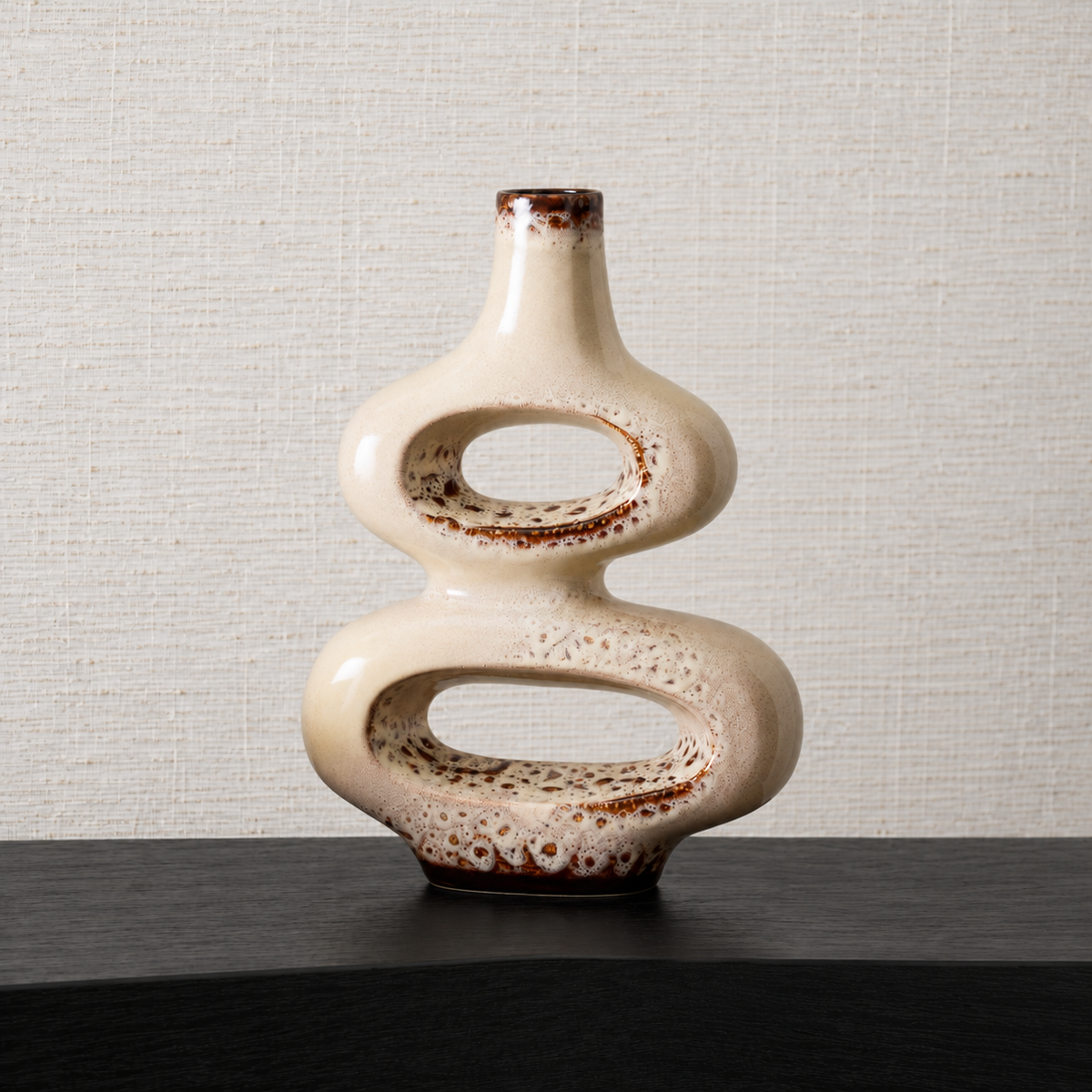 A modern, abstract ceramic vase with a glossy cream-colored glaze and brown speckled accents, displayed on a dark wooden surface against a textured beige wall.
