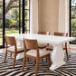 A white rectangular dining table with sculptural pedestal legs and four wooden dining chairs with cream upholstered seats, set on a black and beige zebra-patterned rug in a bright dining room with large windows and beige walls.