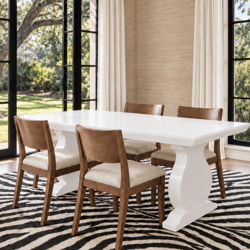 A white rectangular dining table with sculptural pedestal legs and four wooden dining chairs with cream upholstered seats, set on a black and beige zebra-patterned rug in a bright dining room with large windows and beige walls.