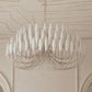 A large white chandelier with multiple elongated teardrop-shaped glass shades and a silver-toned wire frame, hanging from a chain in an elegant cream-colored room with decorative molding on the ceiling and walls.