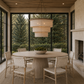 A round light wood dining table with six woven-back chairs in a bright, modern sunroom with large windows, a stone fireplace, and a tiered pendant chandelier.