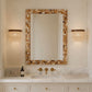 A rectangular wall mirror with a mosaic shell-style frame in beige, brown, and cream tones, mounted above a white marble double vanity with gold fixtures. Two matching cylindrical wall sconces with woven crystal shades flank the mirror on a warm beige wall.