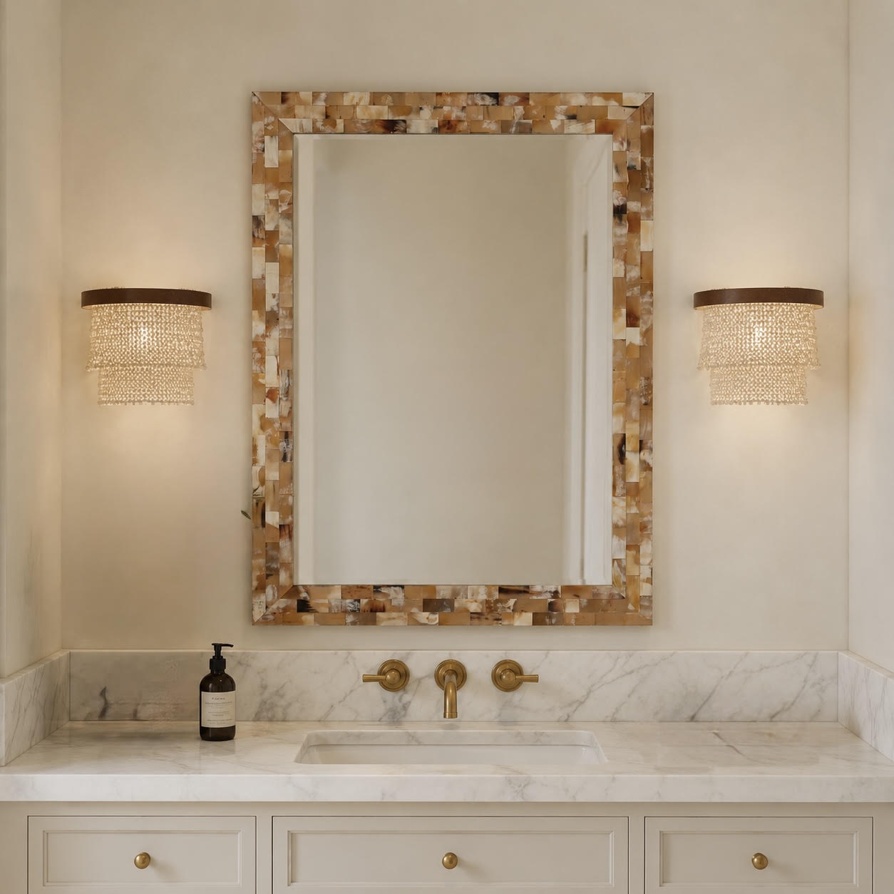 A rectangular wall mirror with a mosaic shell-style frame in beige, brown, and cream tones, mounted above a white marble double vanity with gold fixtures. Two matching cylindrical wall sconces with woven crystal shades flank the mirror on a warm beige wall.
